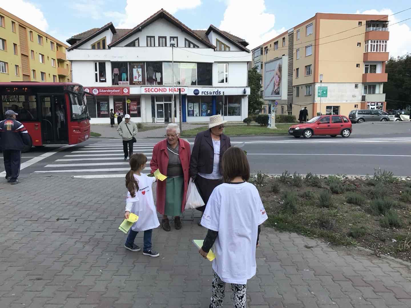 Două voluntare în tricouri albe „Promenada Inimilor” dau pliante galbene unor doamne în vârstă, la trecerea de pietoni din Turda